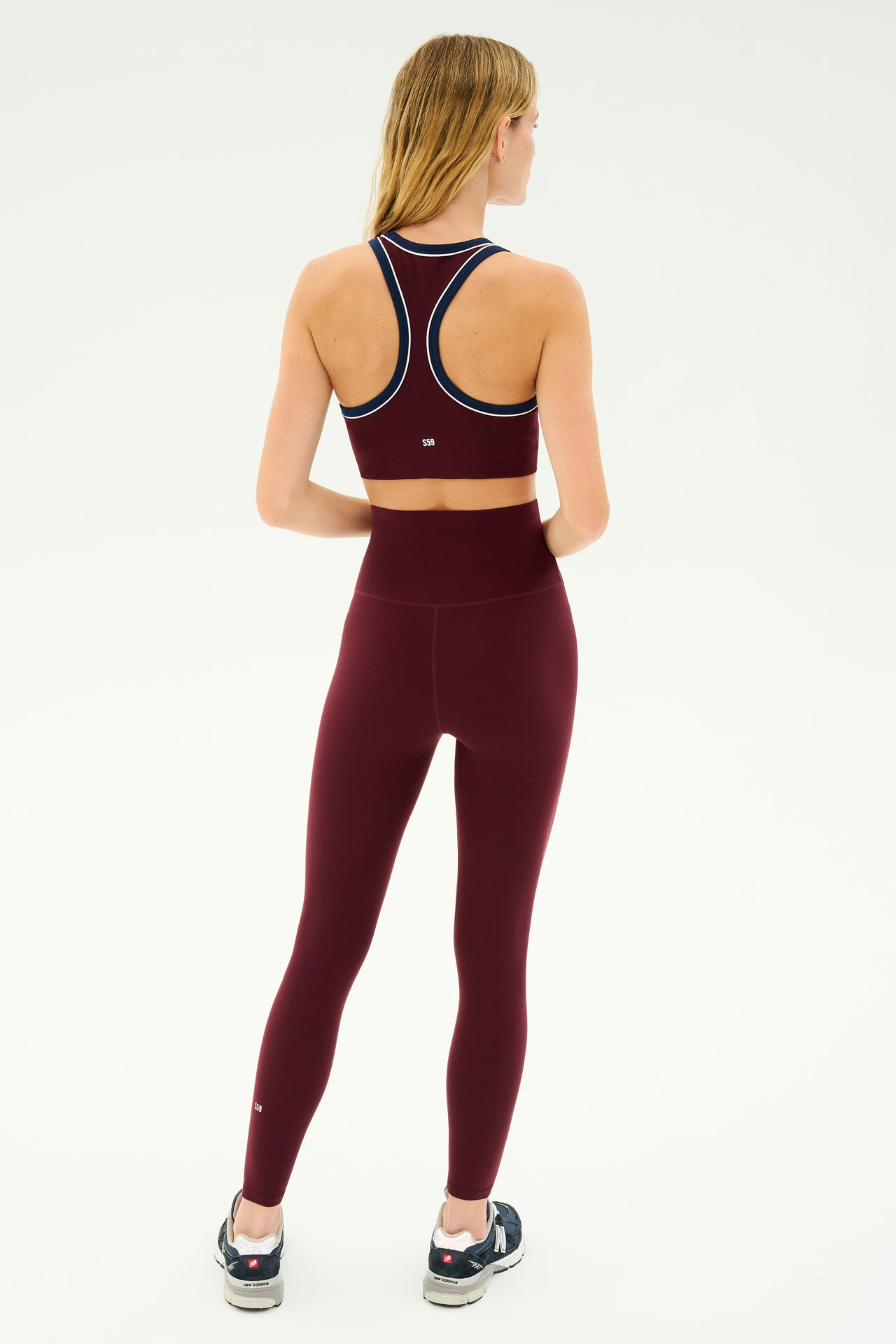 A person is standing with their back turned, wearing a SPLITS59 Dark Cherry Airweight Super High Waist 7/8 leggings and a burgundy sports bra, paired with dark sneakers.