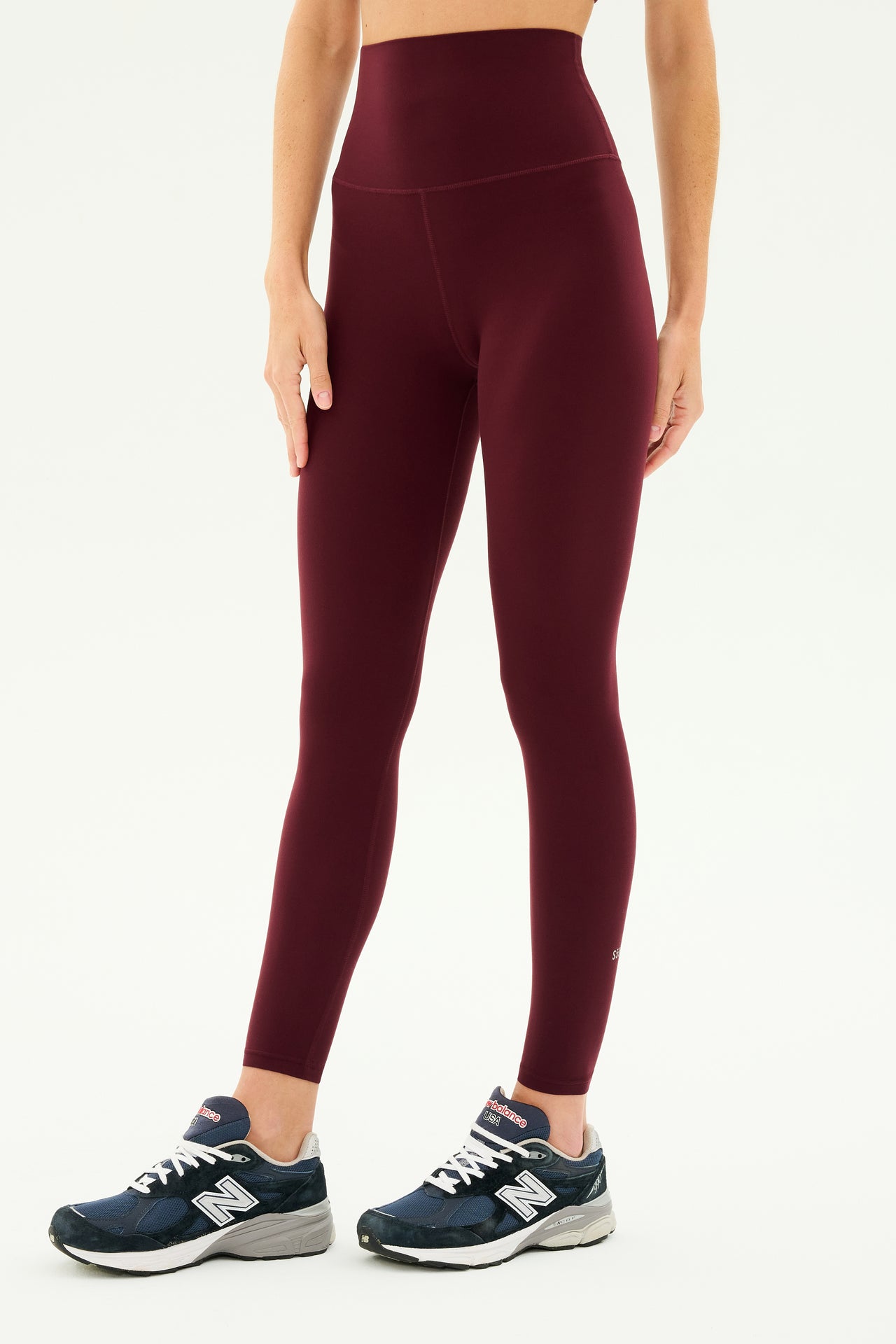 A person wearing the SPLITS59 Airweight Super High Waist 7/8 leggings in Dark Cherry and blue athletic shoes stands against a light background, ready for hot yoga.