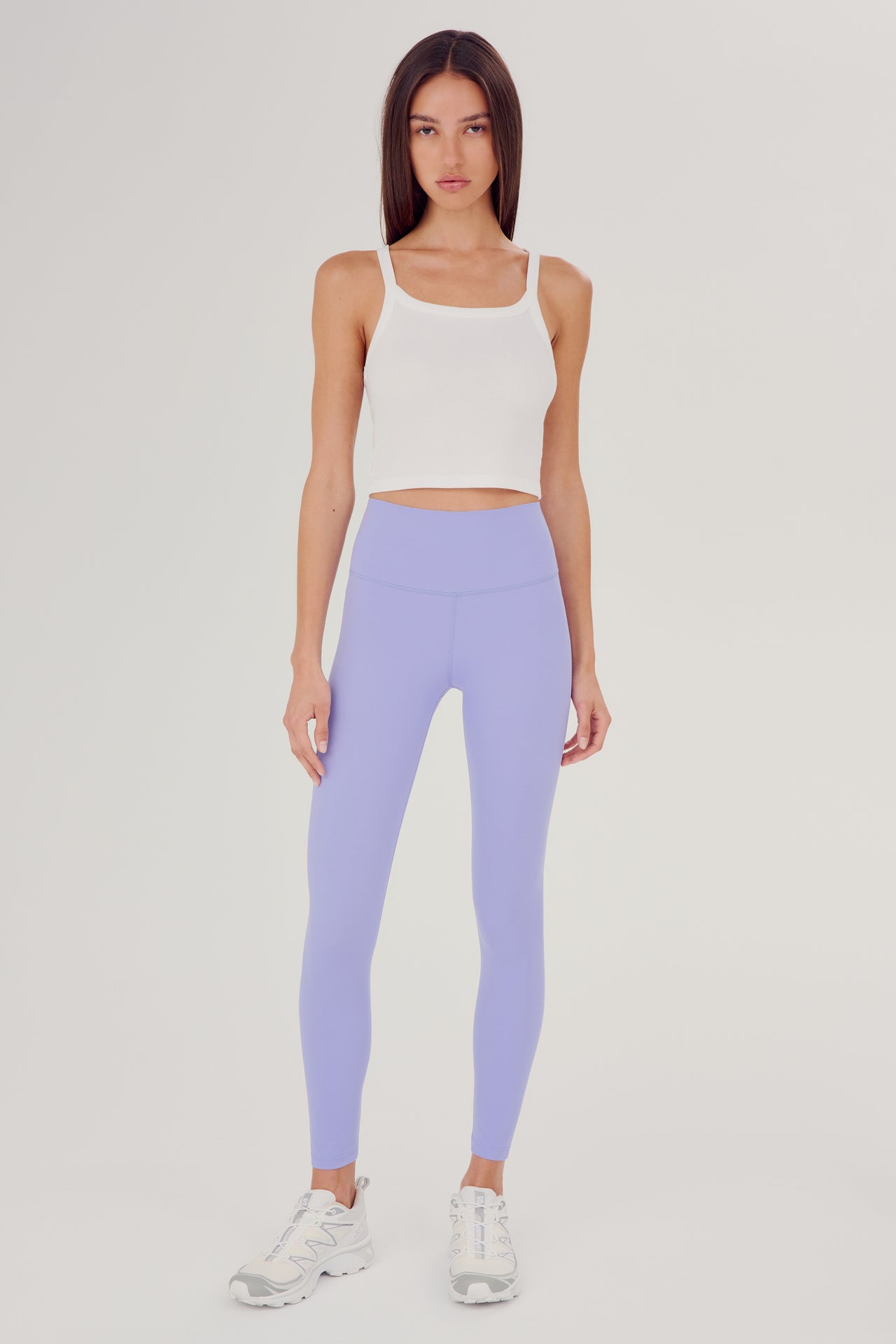 Front full view of woman with dark straight hair wearing light purple high waist  leggings with white cropped tank and white shoes