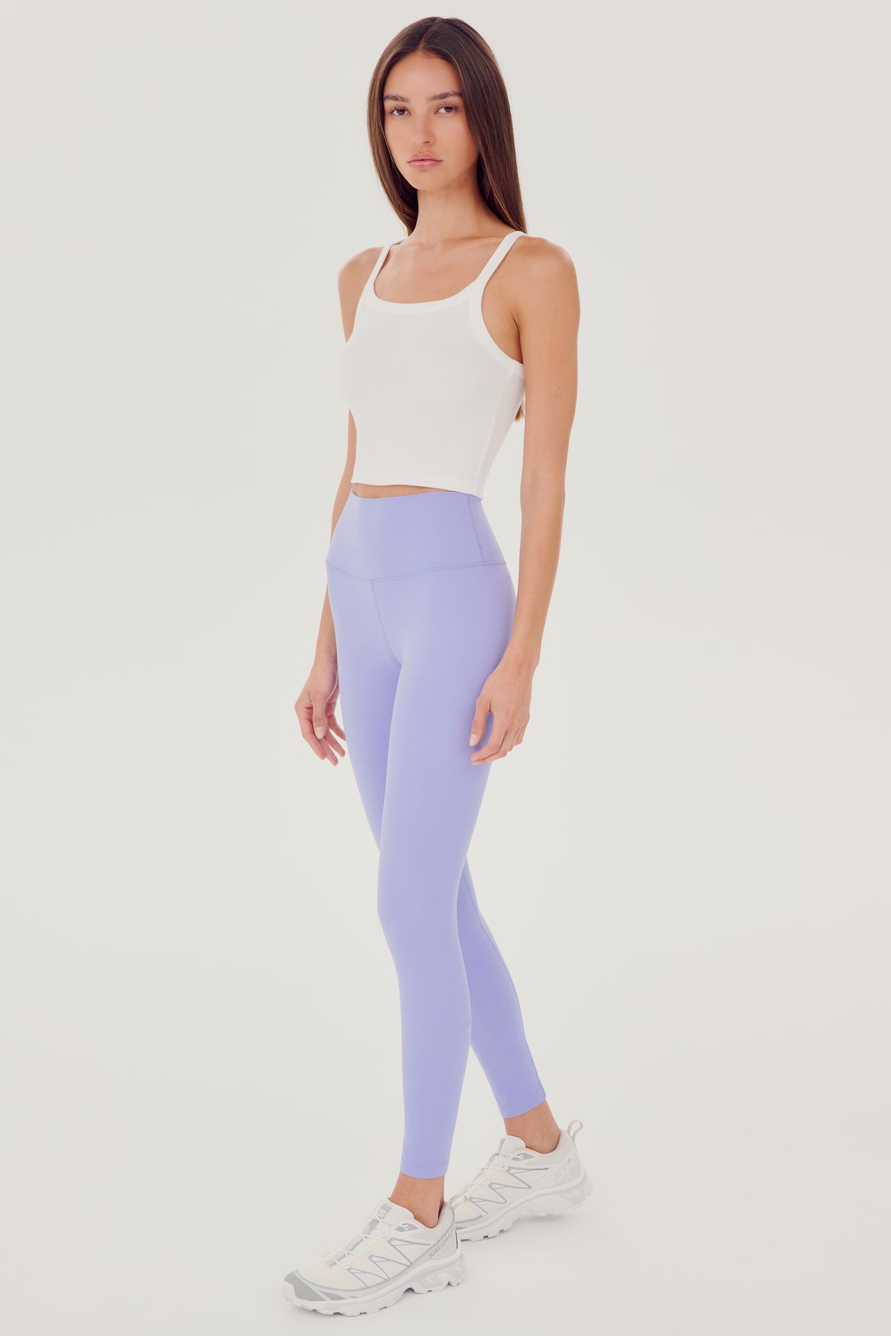 Front full view of woman with dark straight hair wearing light purple high waist leggings with white cropped tank and white shoes