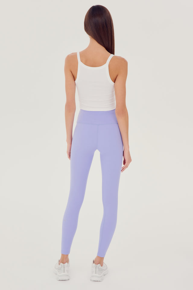 Back full view of woman with dark straight hair wearing light purple high waist  leggings with white cropped tank and white shoes