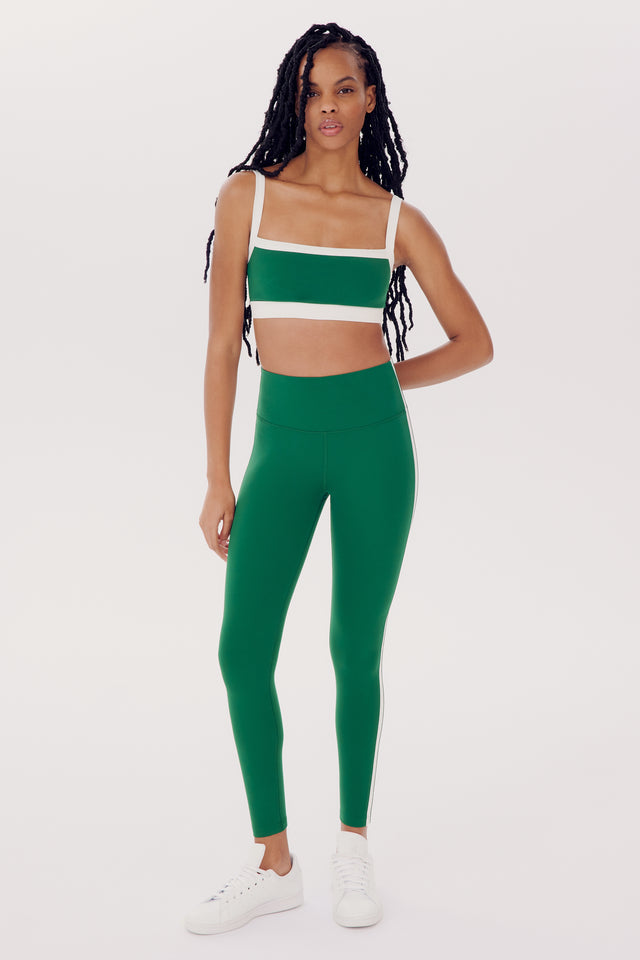 A woman wearing a green sports bra and SPLITS59 Clare High Waist Rigor 7/8 - Arugula/White leggings stands confidently against a white background.
