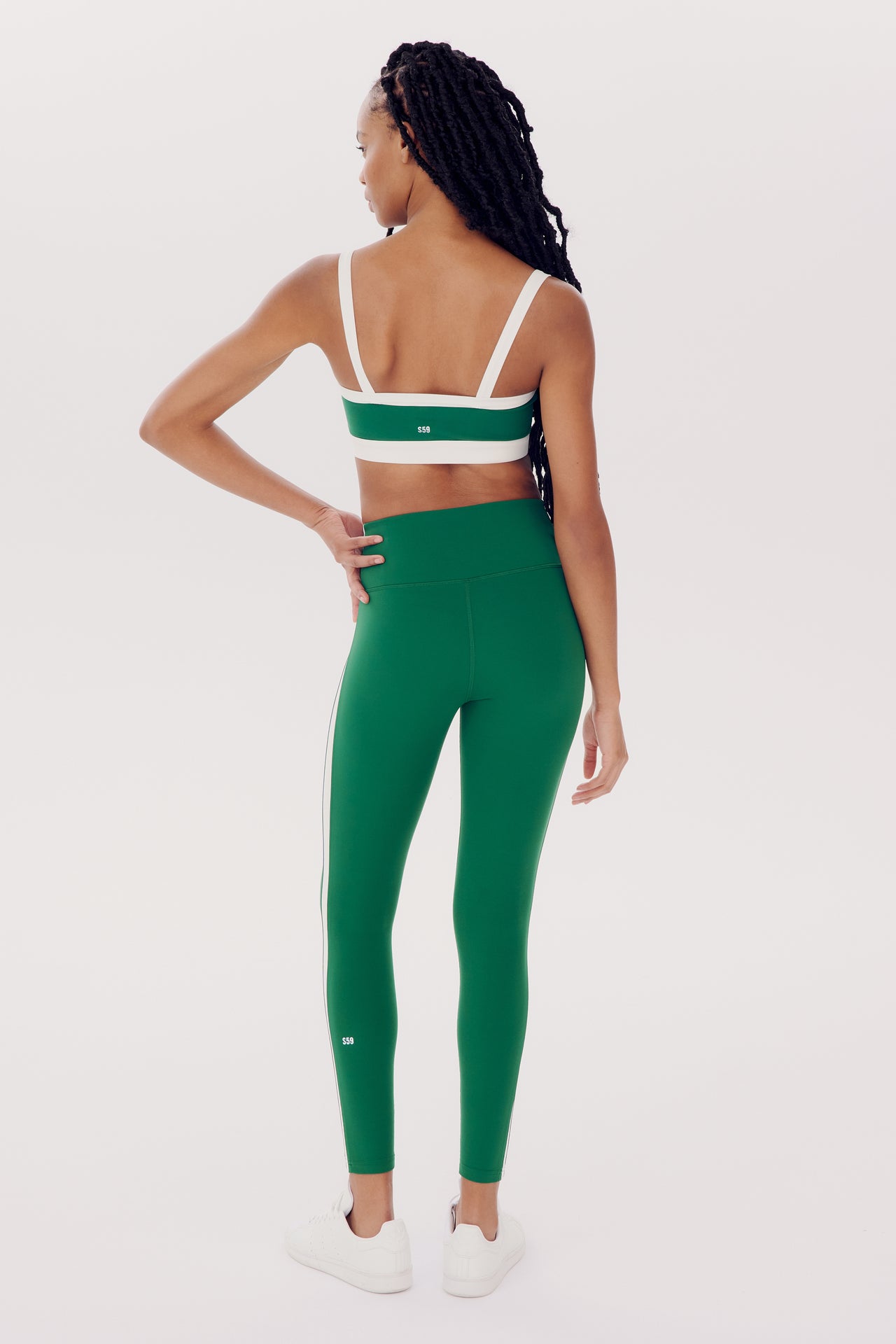 Woman in Clare High Waist Rigor 7/8 - Arugula/White and green sports bra standing with her back to the camera, looking over her shoulder, against a white background by SPLITS59.