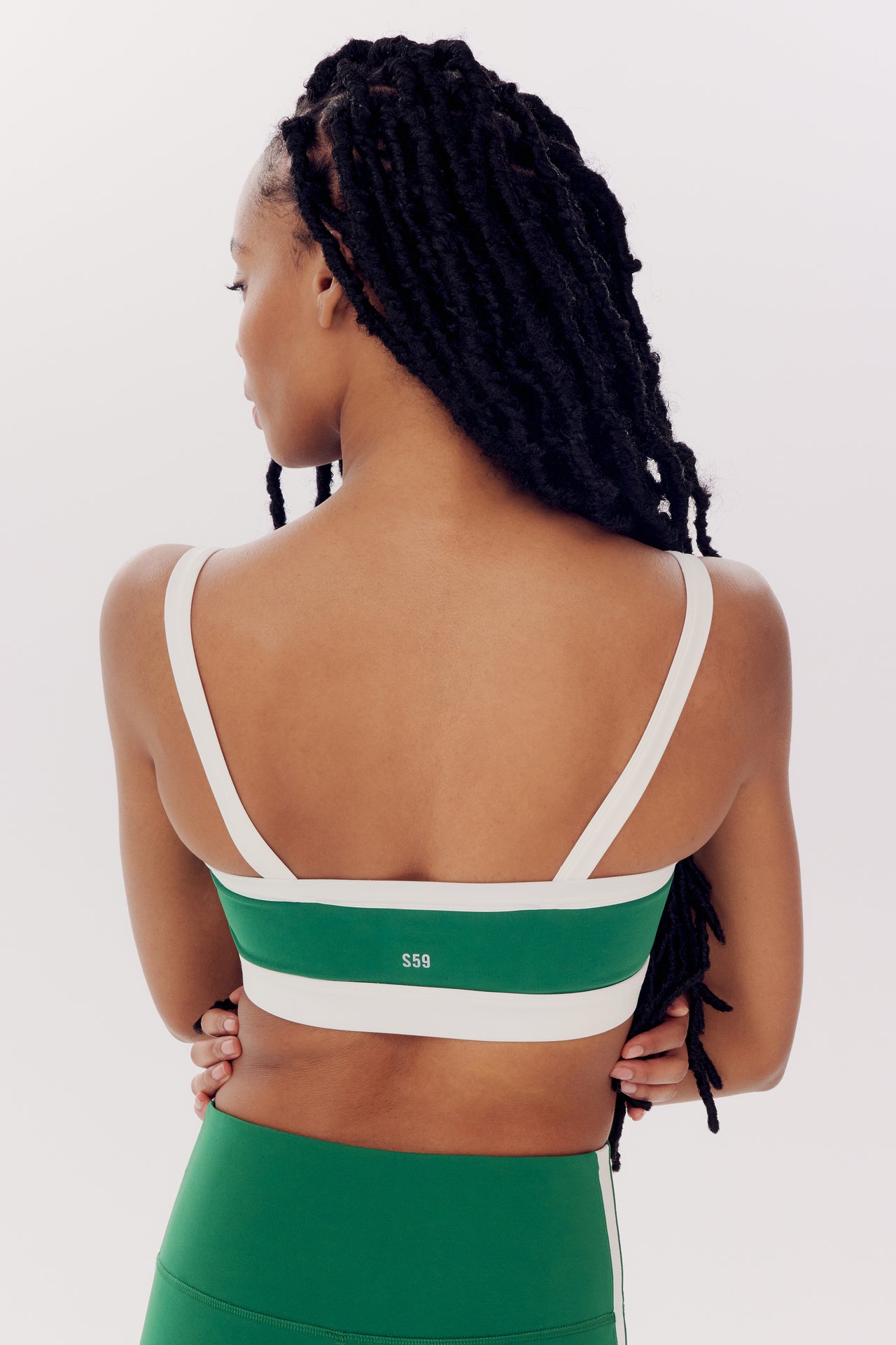 Rear view of a woman with braided hair wearing a white and green SPLITS59 Monah Rigor Bra in Arugula/White, standing against a white background.