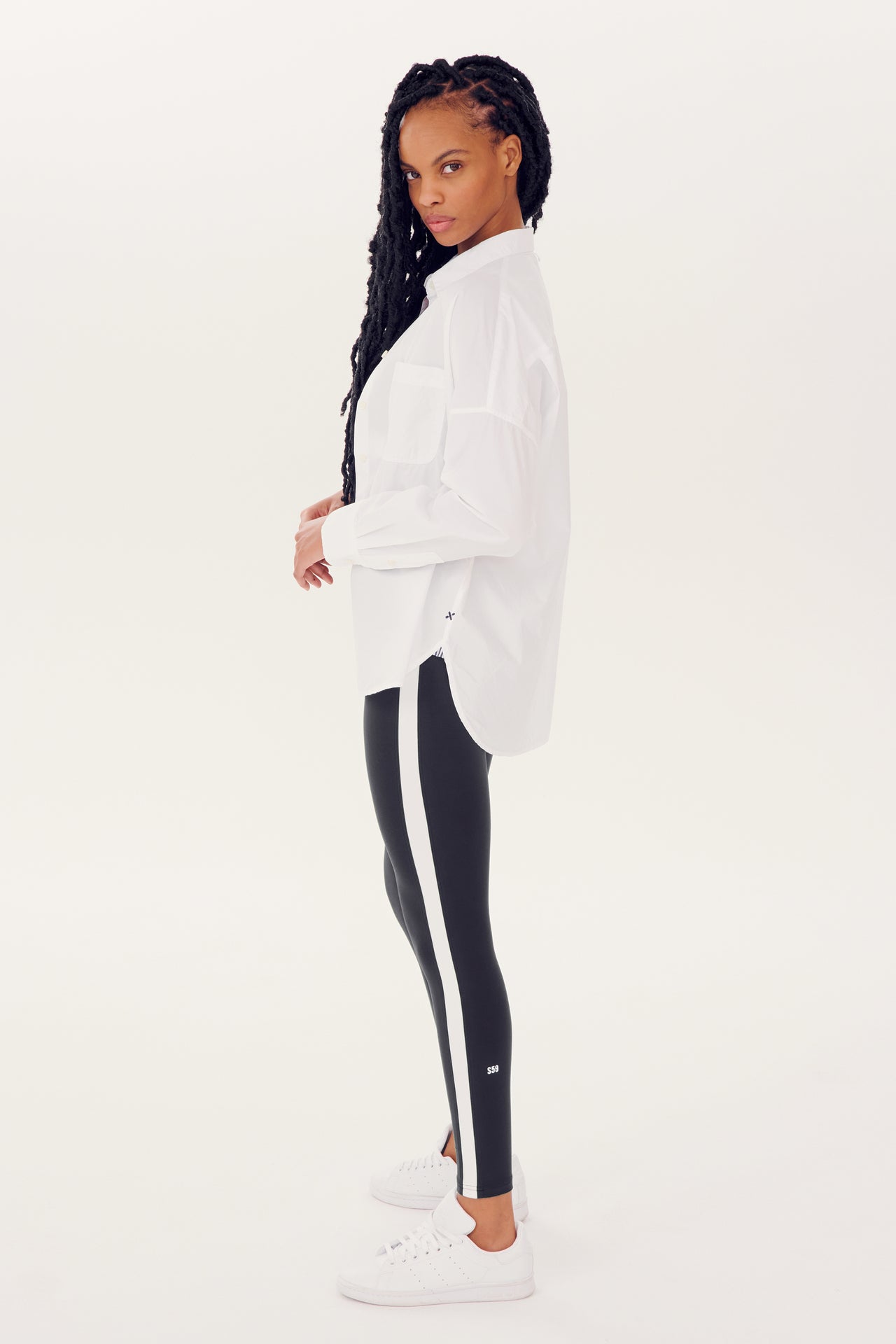 Woman posing in profile wearing a white Alex Mill x Splits59 Jo Shirt, black leggings with a white stripe from Splits59 looks, and white sneakers.