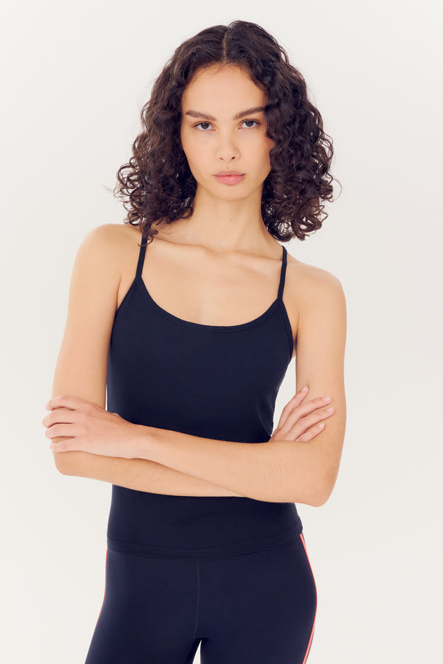 A young woman with curly hair wearing a SPLITS59 Airweight Tank in Indigo and leggings stands with her arms crossed, looking directly at the camera against a white background.
