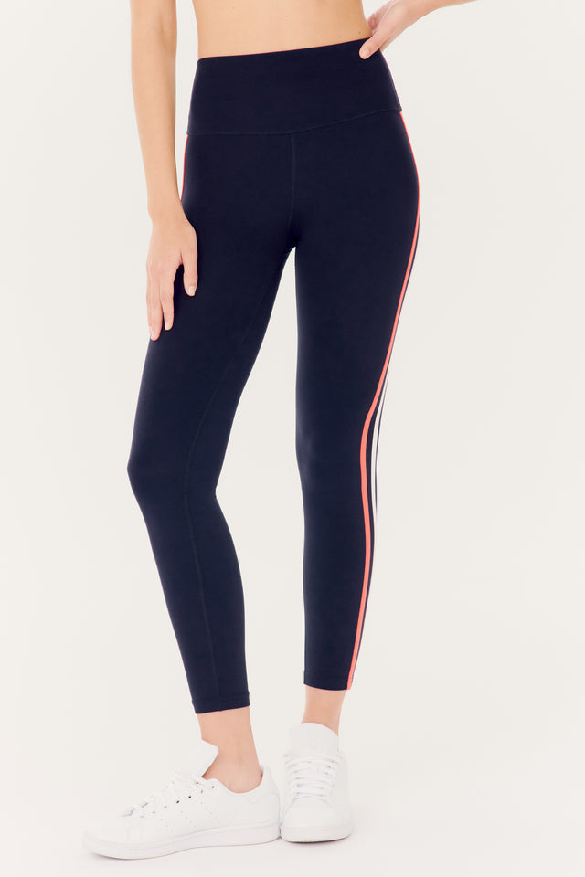 Woman wearing SPLITS59 Ella High Waist Airweight 7/8 leggings in Indigo/Melon with a side stripe and white sneakers.