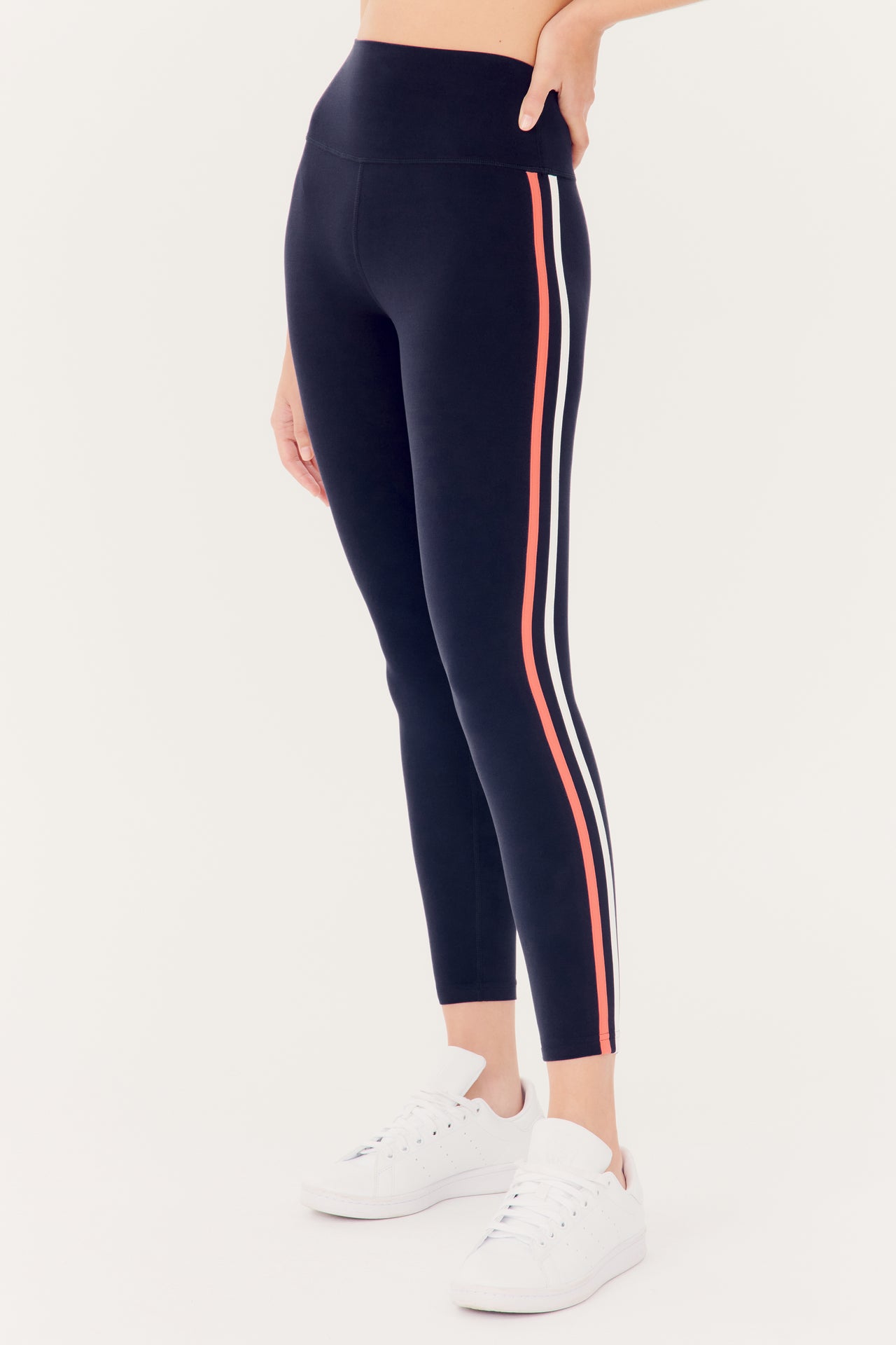 A person standing sideways wearing SPLITS59 Ella High Waist Airweight 7/8 leggings in Indigo/Melon with a side stripe and white sneakers.