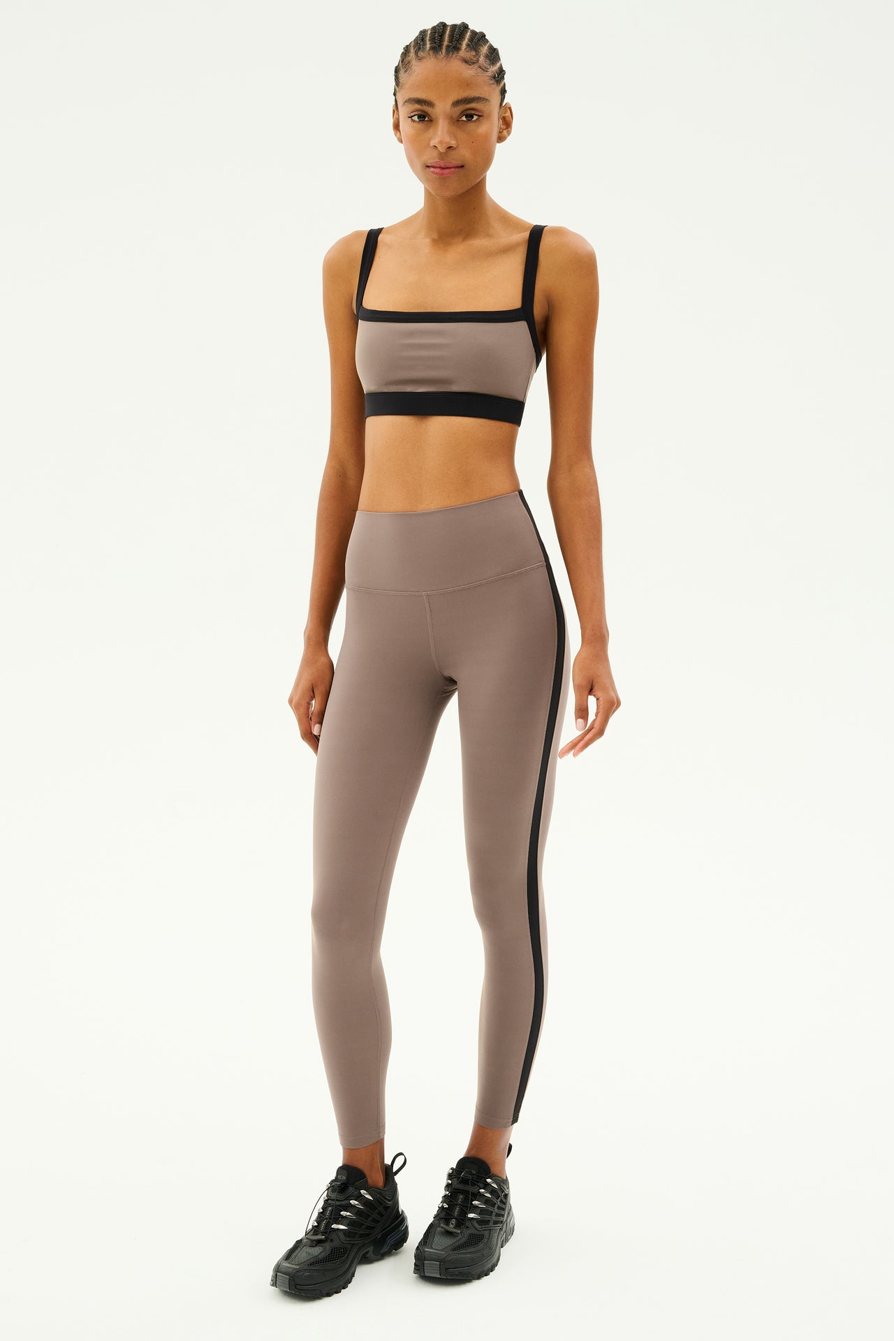 Clad in the Monah Rigor Bra in Lentil/Black by SPLITS59, a person with braids stands confidently against a plain backdrop, ready for gym workouts.