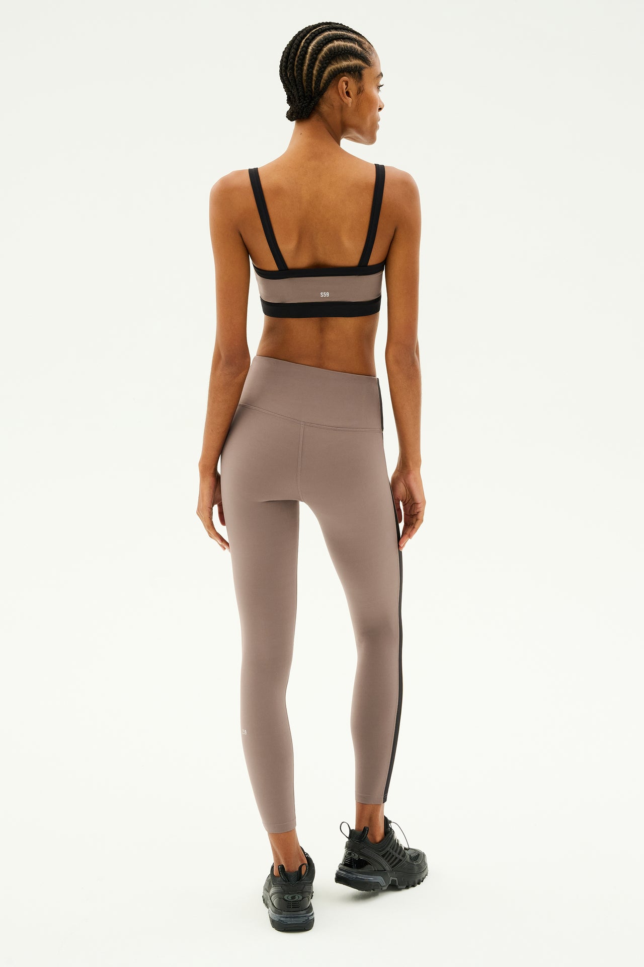 Clad in the Monah Rigor Bra from SPLITS59 and matching leggings, a person stands with their back turned, prepared for exercise in black sneakers against a plain backdrop.