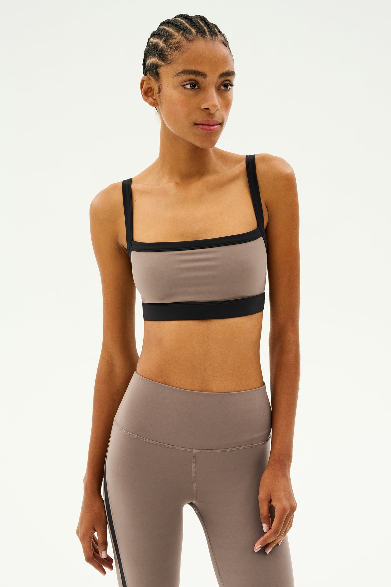 A person wearing a beige and black athletic outfit, featuring the stylish SPLITS59 Monah Rigor Bra in Lentil/Black, stands against a plain background.