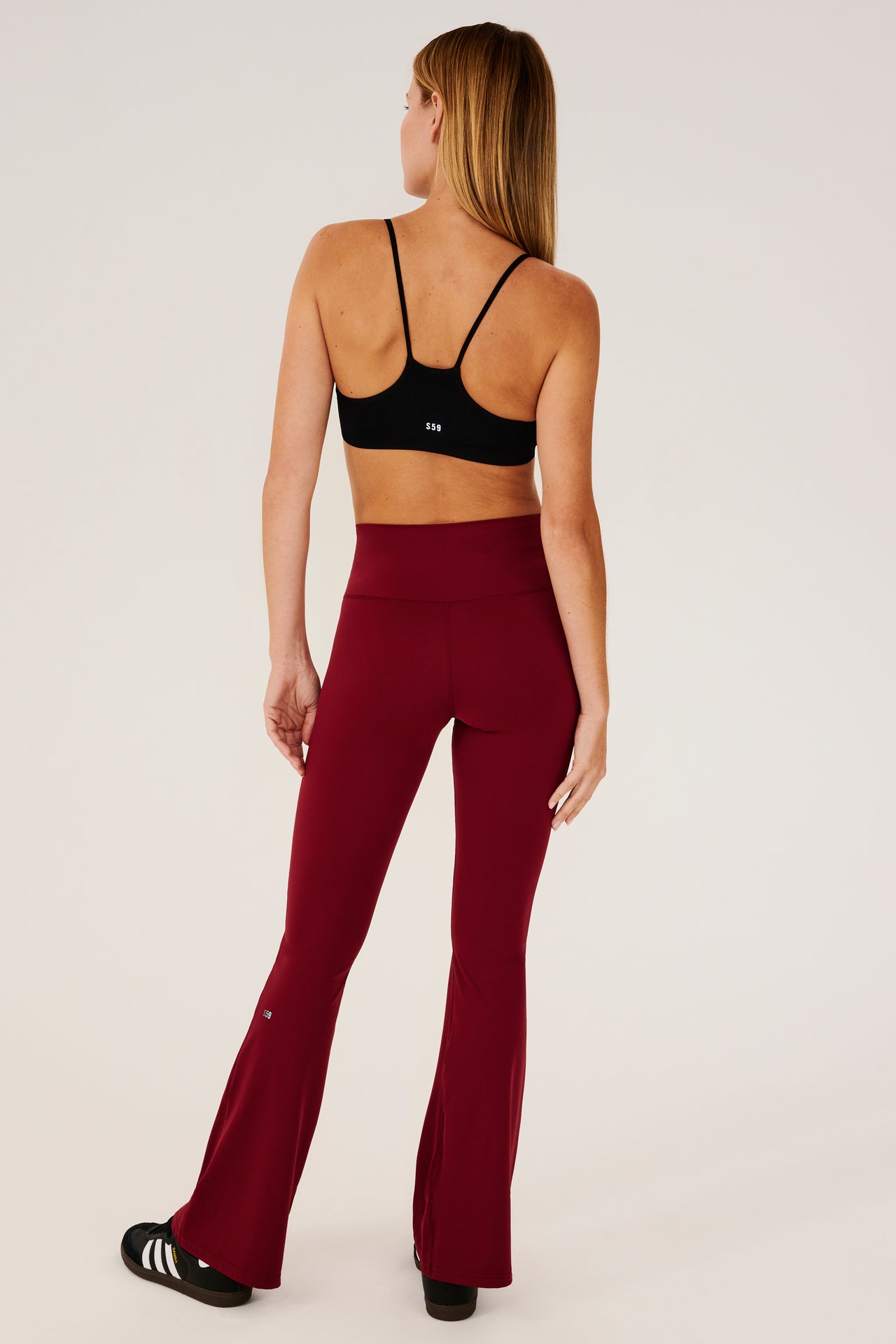 Full back view of woman with straight blonde hair wearing a dark red high waist below ankle length legging with wide flared bottoms with black S59 logo on back of left calf and black racerback bra with spaghetti straps and white S59 logo. Paired with black shoes with white stripes. 