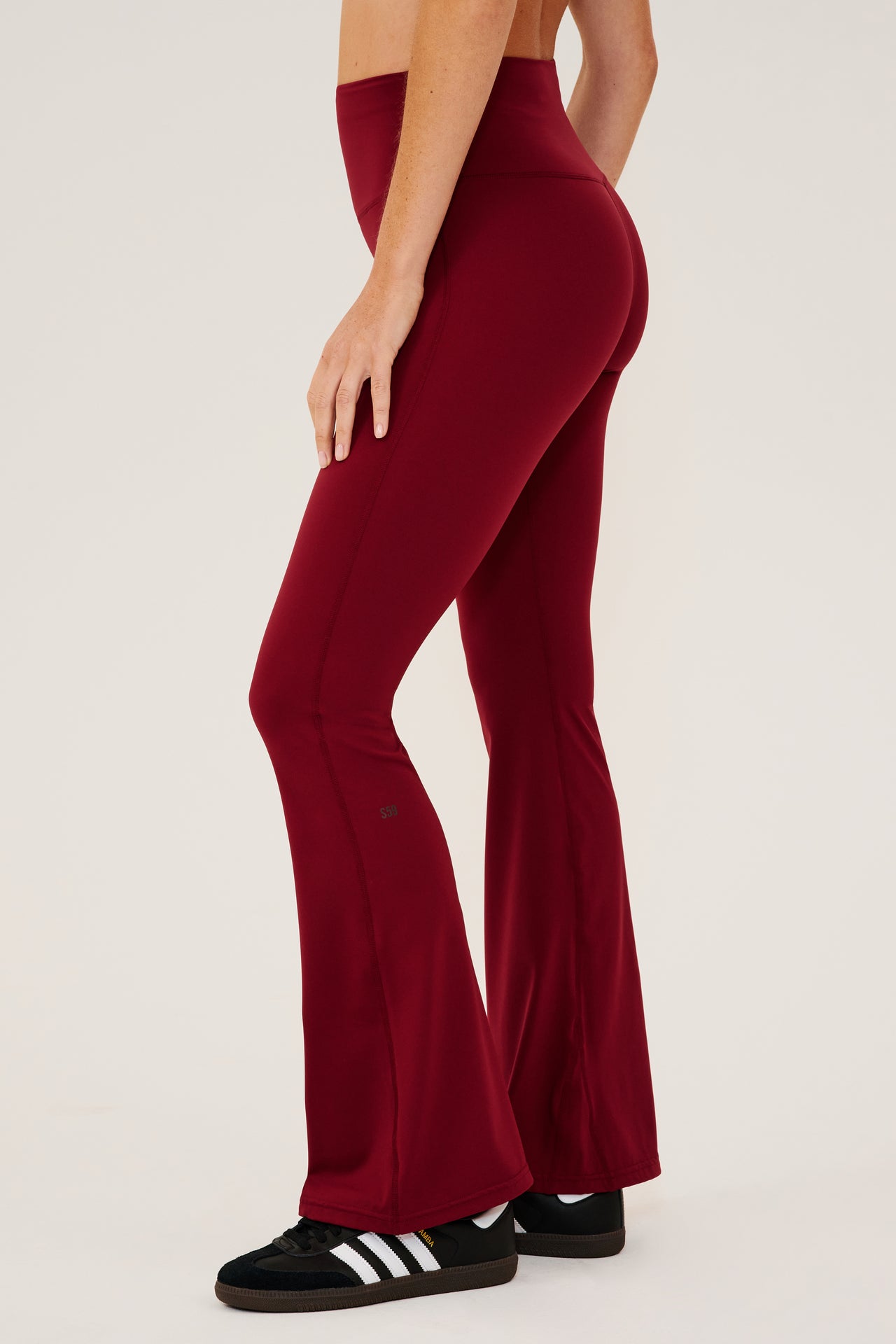 Side view of dark red high waist below  ankle length legging with wide flared bottoms with black S59 logo on back of left calf. Paired with black shoes with white stripes.