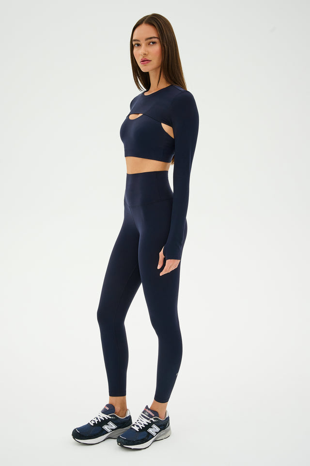 Against a plain backdrop, a person stands poised like a dancer in a dark athletic outfit and sneakers, their posture suggesting readiness for an Amelia Airweight Lite Shrug in Indigo from SPLITS59.