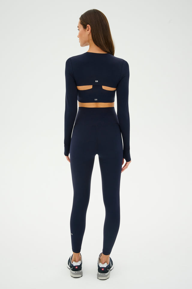 A woman exudes grace in an SPLITS59 Amelia Airweight Lite Shrug - Indigo, her workout outfit featuring back cutouts. She stands confidently, ready for yoga or dance in black sneakers against a plain background.