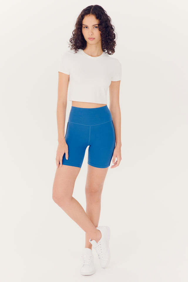 Woman posing in SPLITS59 Airweight S/S Crop - White top and blue spandex shorts with white sneakers.