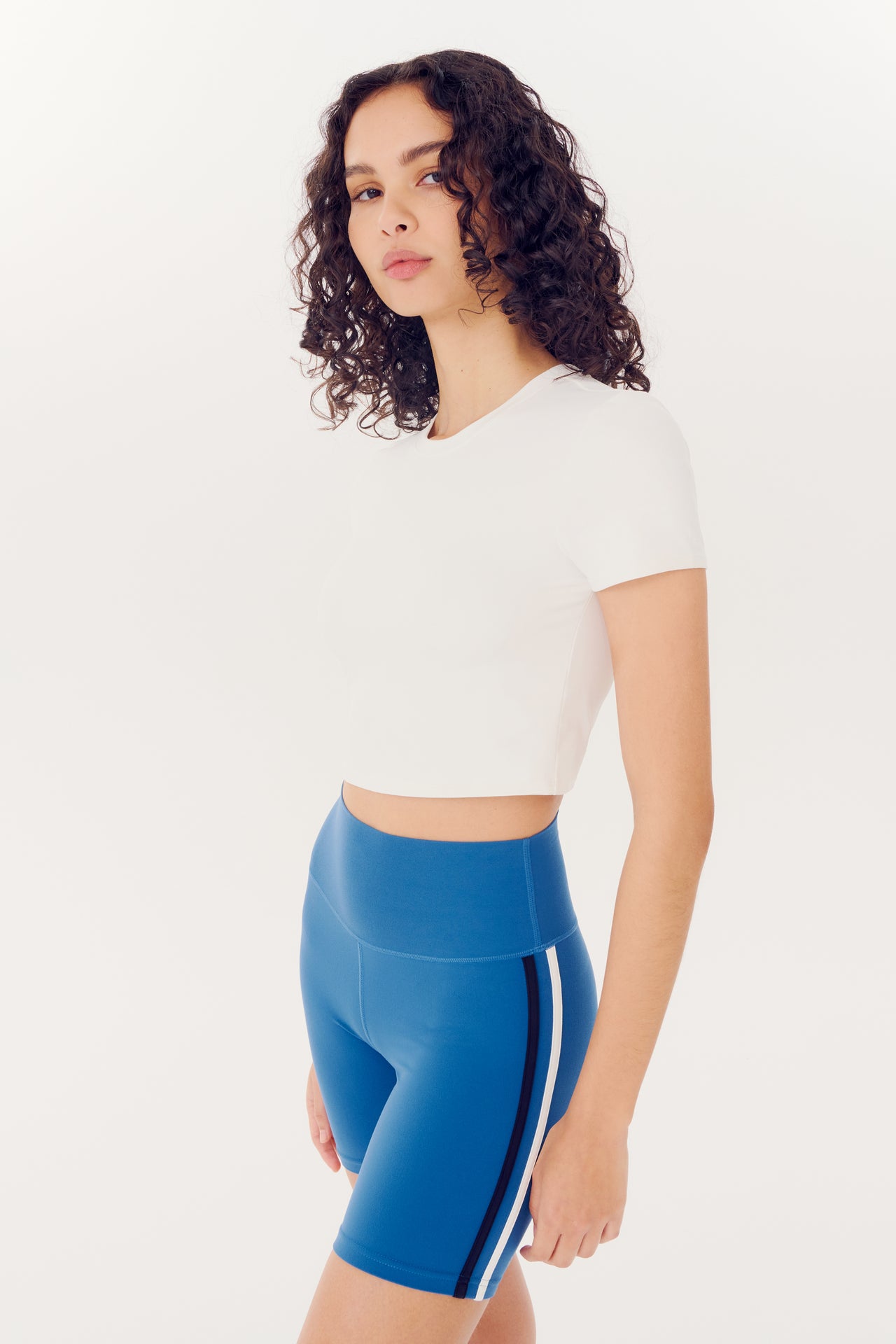 A woman in a SPLITS59 white Airweight S/S Crop top and blue nylon athletic shorts with a white stripe poses against a white background.