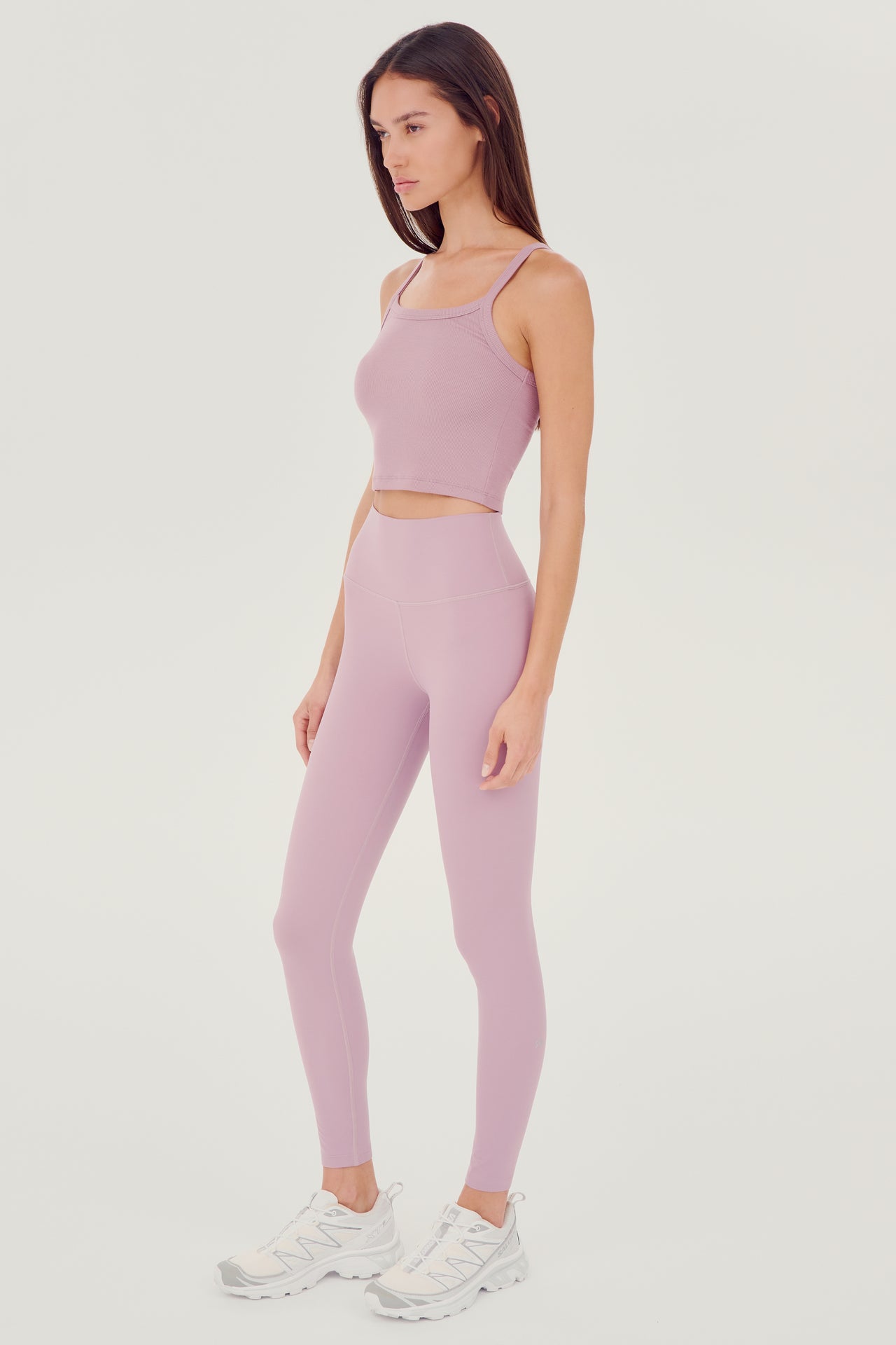 Full front side view of woman with straight dark hair wearing light pink high waist legging and light pink bra tank paired with white shoes