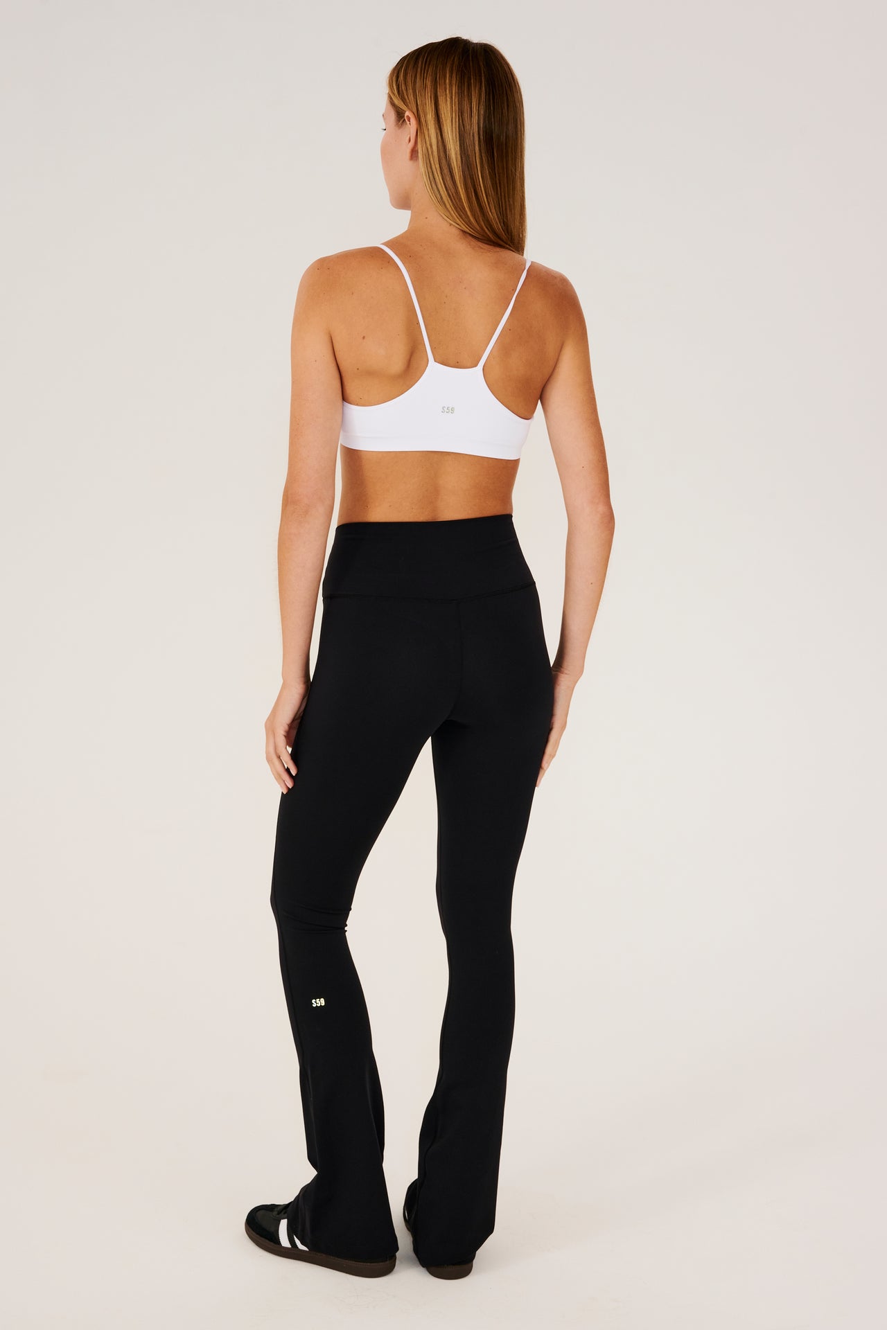 Full back view of woman with straight blonde hair wearing a black high waist below ankle length legging with wide flared bottoms with gray S59 logo on back of left calf and white racerback bra with spaghetti straps. Paired with black shoes with white stripes.
