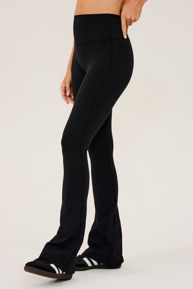 Front side view of woman wearing a black high waist below ankle length legging with wide flared bottoms with gray S59 logo on back of left calf. Paired with black shoes with white stripes.