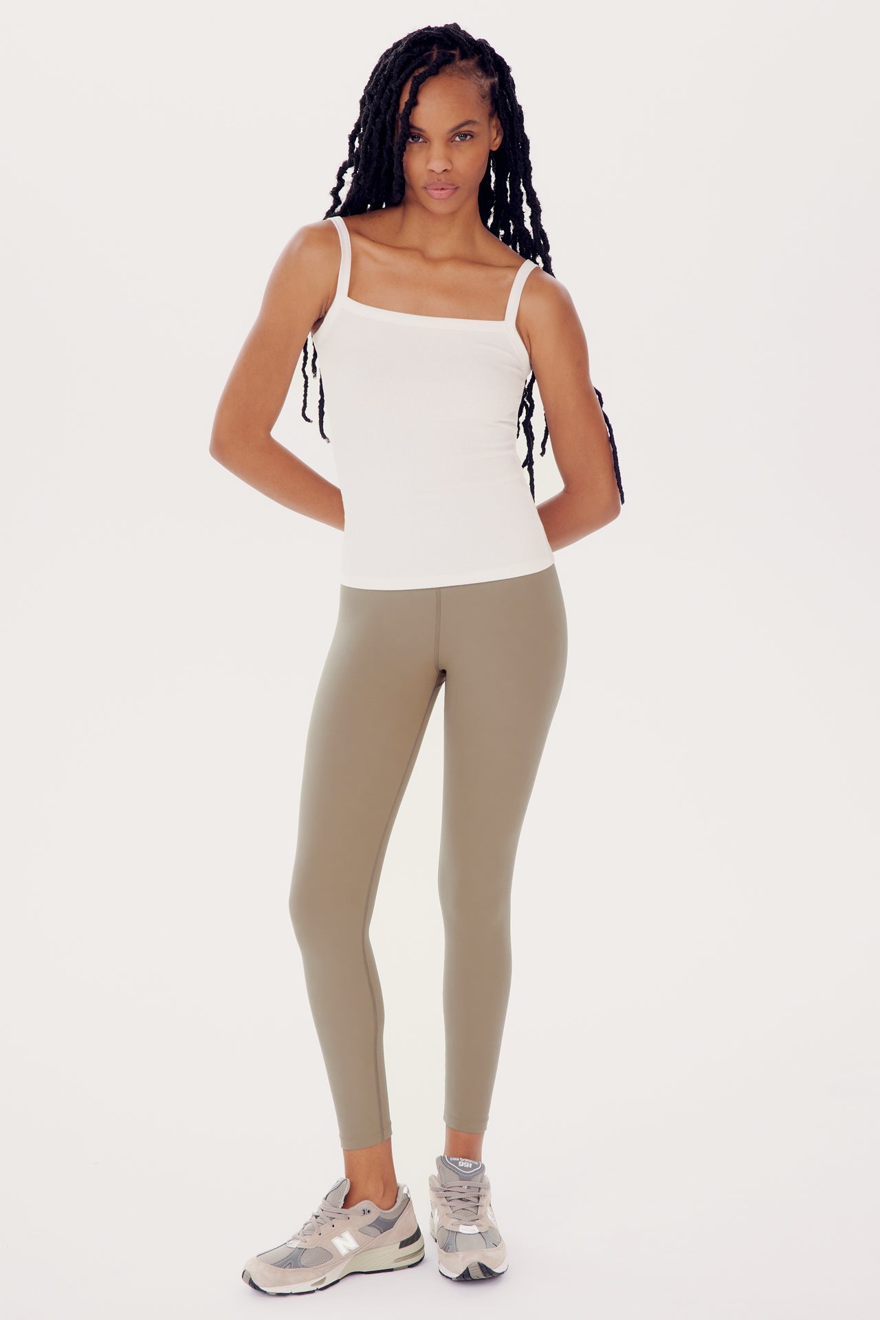 A woman in a white tank top and olive green Sprint High Waist Rigor 7/8 - Latte leggings stands with her hands at her sides, wearing silver sneakers, against a white background.