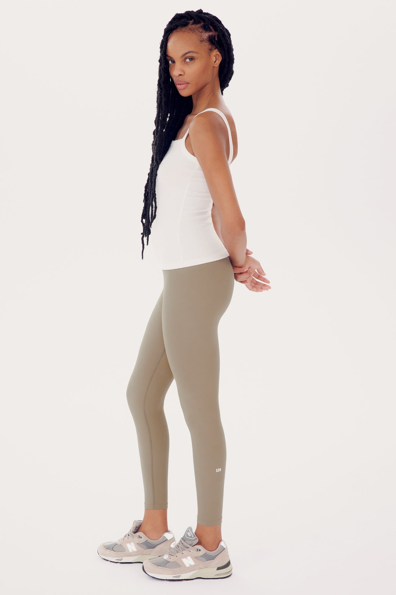 A woman in a white tank top and SPLITS59 Sprint High Waist Rigor 7/8 - Latte leggings stands sideways, looking at the camera, with long braided hair and sneakers.