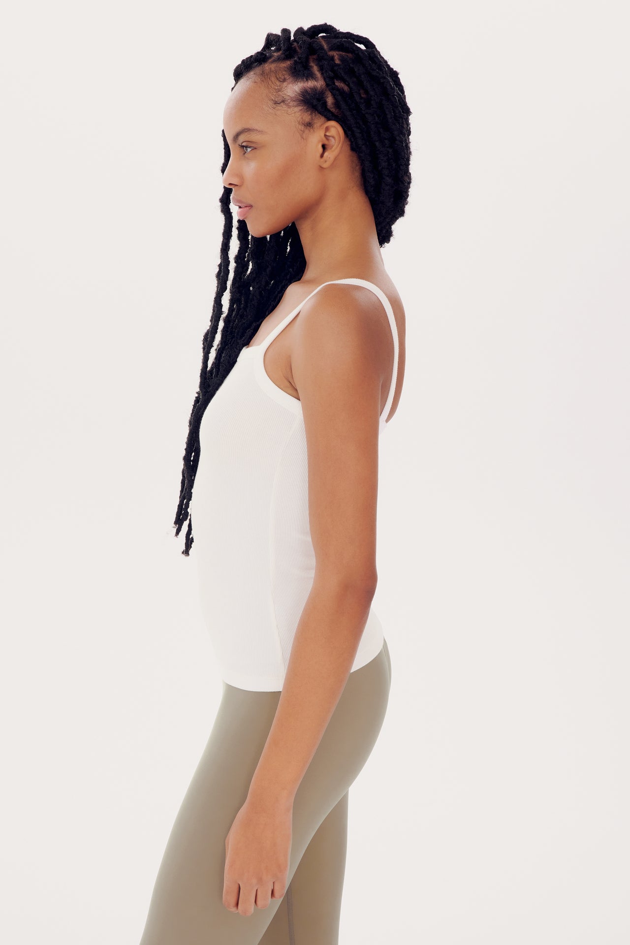 Profile view of a woman with braided hair, wearing an SPLITS59 Romy Rib Tank in White and green leggings, standing against a plain background.