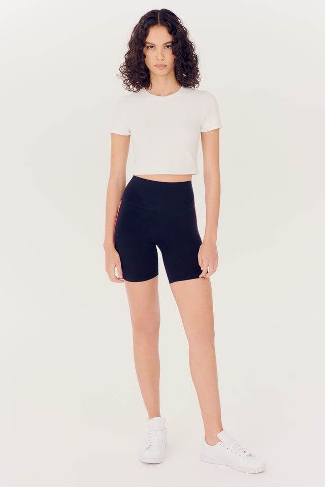 A woman standing against a plain background, wearing a white crop top, the SPLITS59 Ella High Waist Airweight Short in Indigo/Melon, and white sneakers.