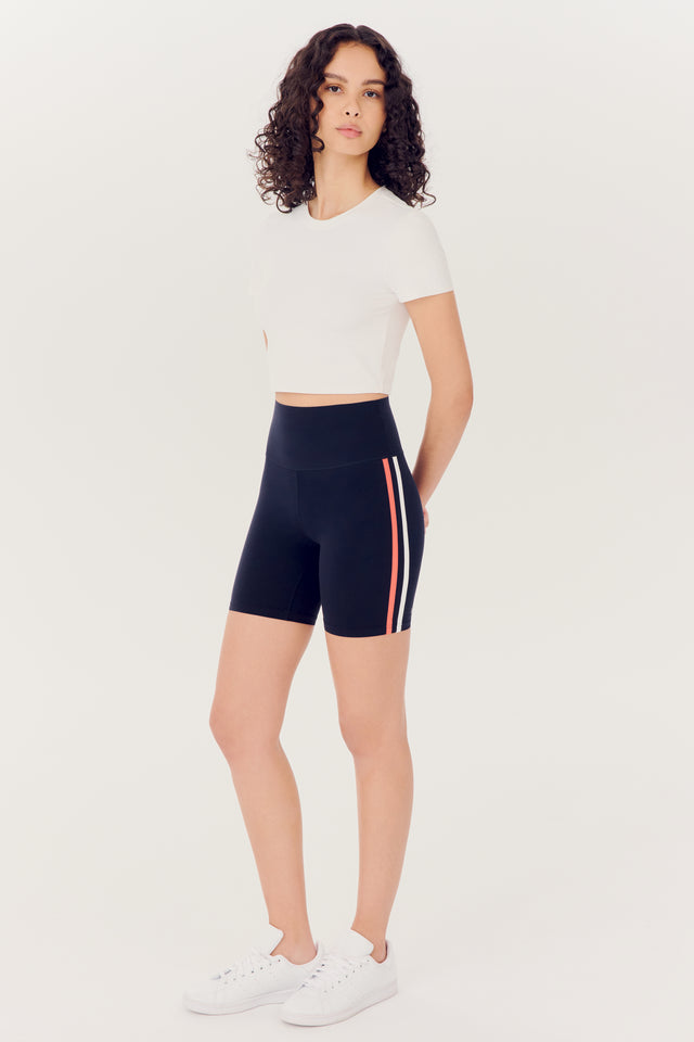 Woman in a white top and black SPLITS59 Ella High Waist Airweight Short with a stripe detail standing against a white background.