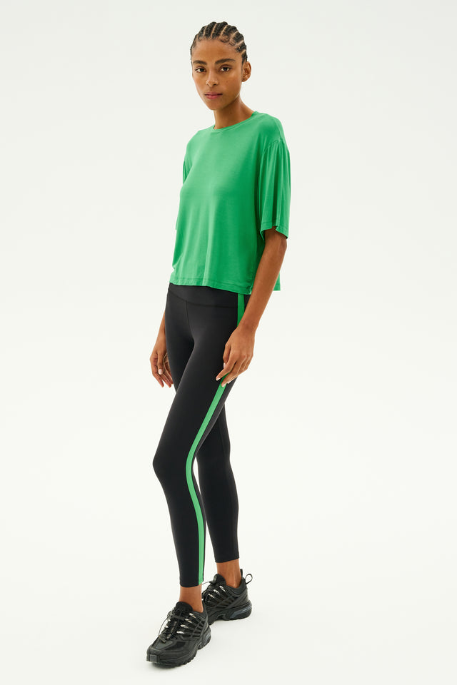 A person wearing the SPLITS59 Djuna Oversized Jersey Tee in Grass stands against a plain background, radiating the energy of gym workouts while pairing it with black leggings featuring green stripes.