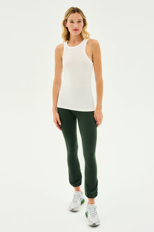 A woman stands against a plain background, wearing a white sleeveless top, the SPLITS59 Icon High Waist Supplex Leggings in Military green, and gray sneakers.