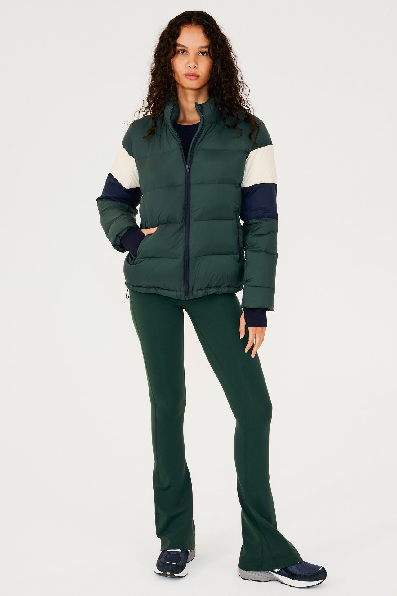 Full front view of girl wearing dark green puffer jacket with blue and white stripes on arms and sleeve cuffs with thumb holes and dark green flared leggings with dark blue shoes