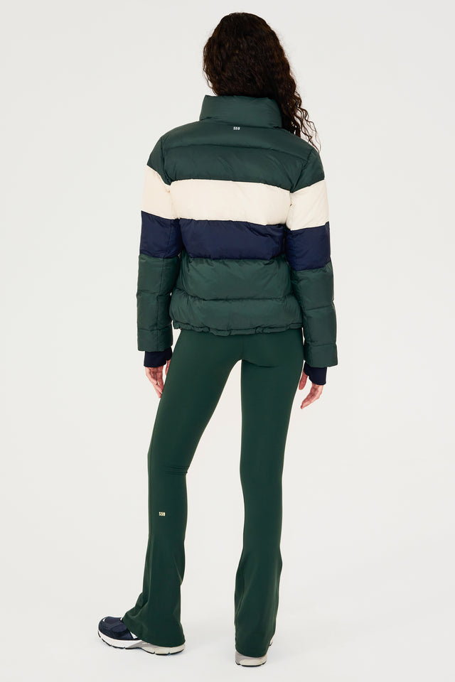 Full side view of girl wearing dark green puffer jacket with blue and white stripes on arms and sleeve cuffs with thumb holes and dark green flared leggings with dark blue shoes