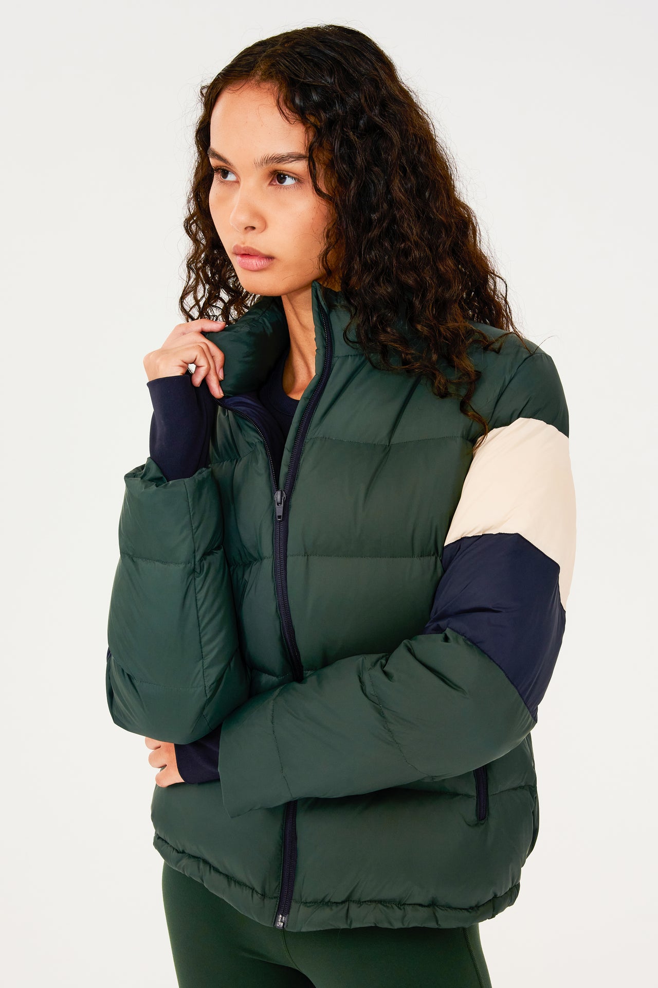Side view of girl wearing dark green puffer jacket with blue and white stripes on arms and sleeve cuffs with thumb holes