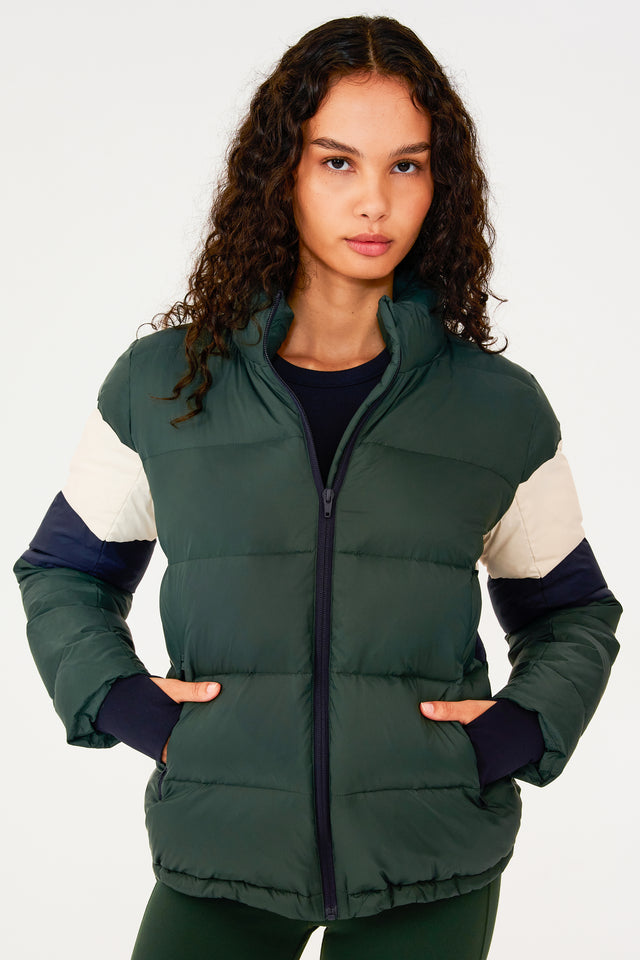 Front view of girl wearing dark green puffer jacket with blue and white stripes on arms and sleeve cuffs with thumb holes