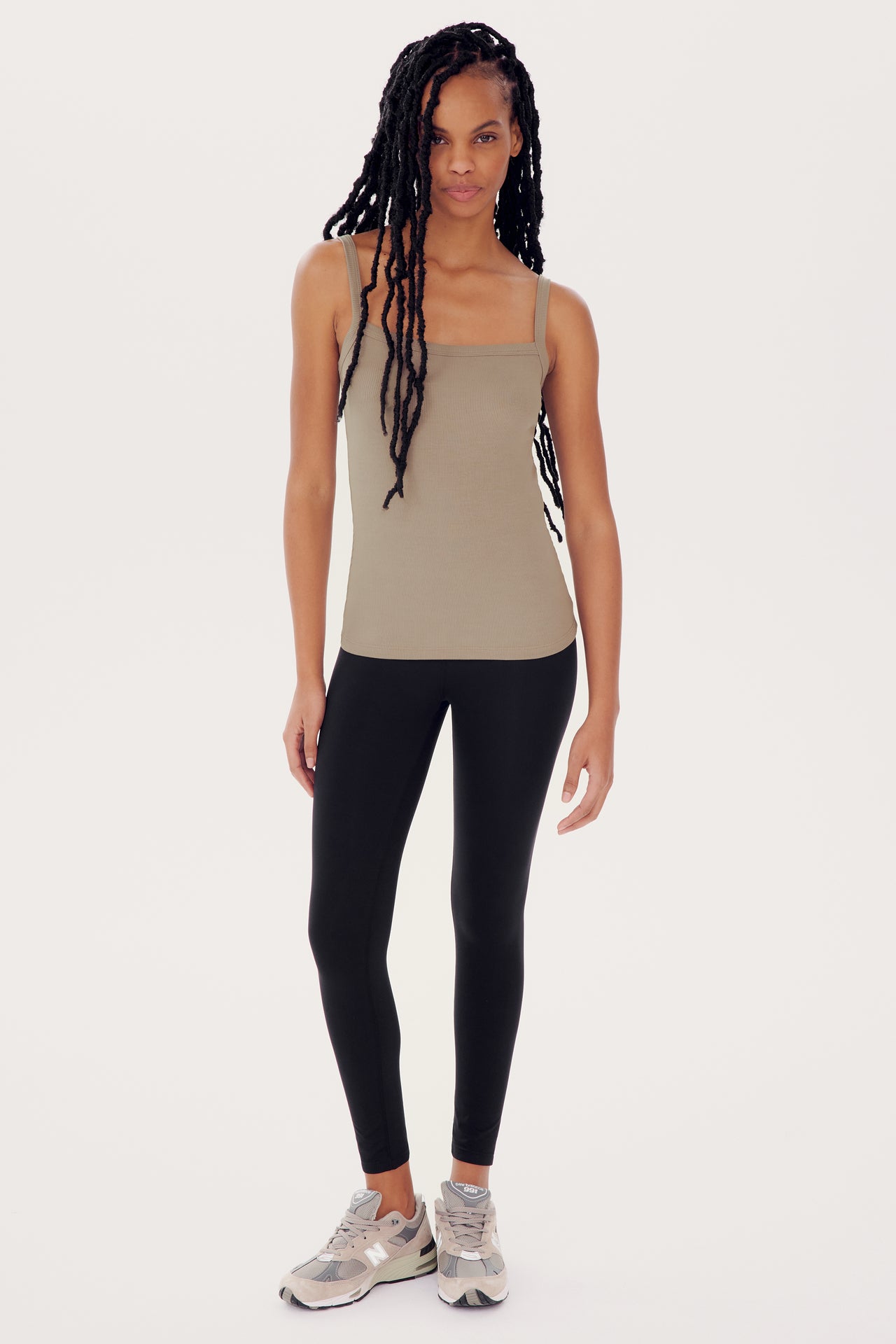 A woman wearing a Romy Rib Tank in Latte by SPLITS59 with a square neckline and black leggings stands confidently against a white background.