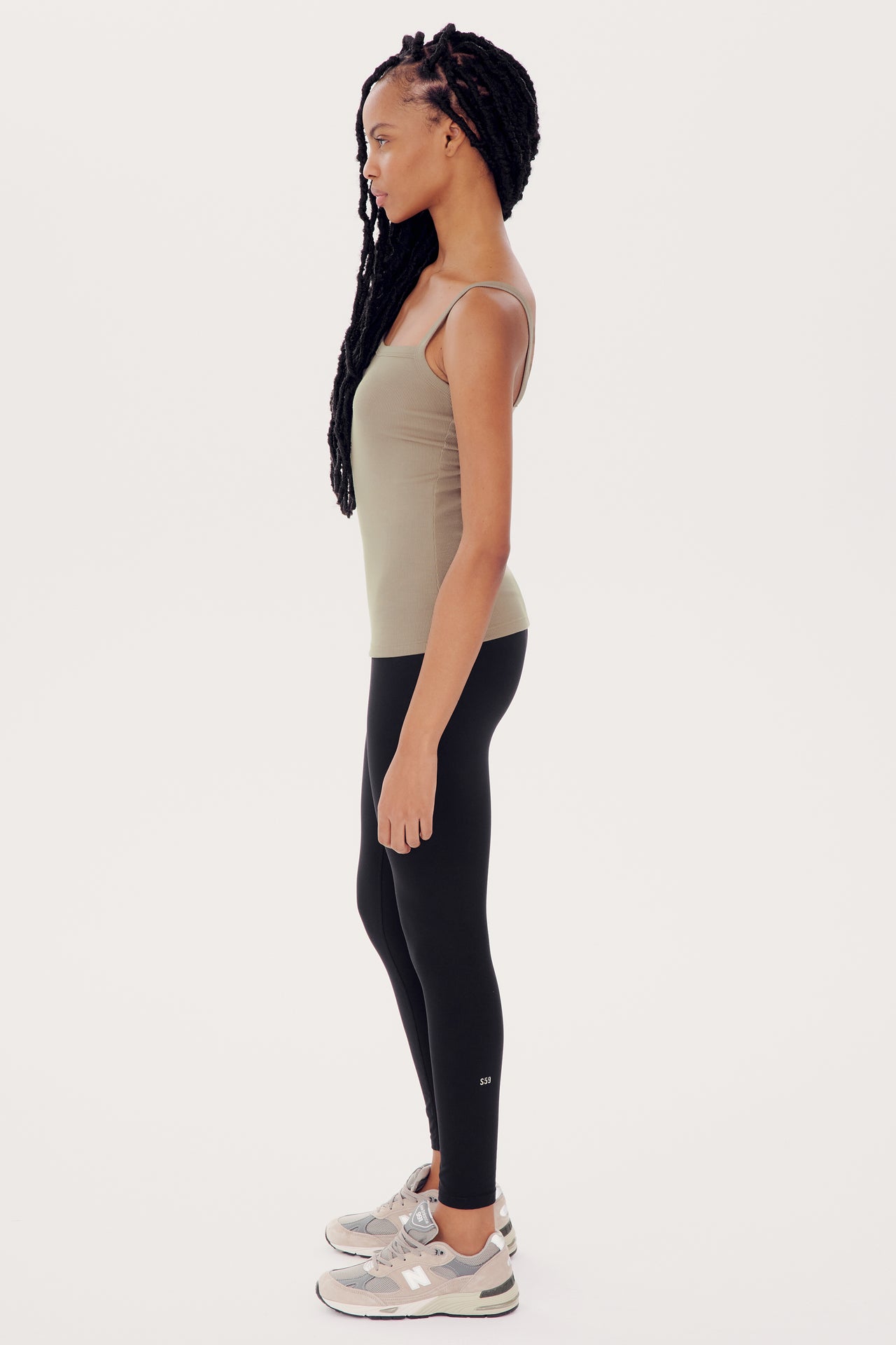 A woman in a Romy Rib Tank in Latte by SPLITS59 with a square neckline and black leggings standing sideways, with her hands slightly touching her thighs, against a white background.