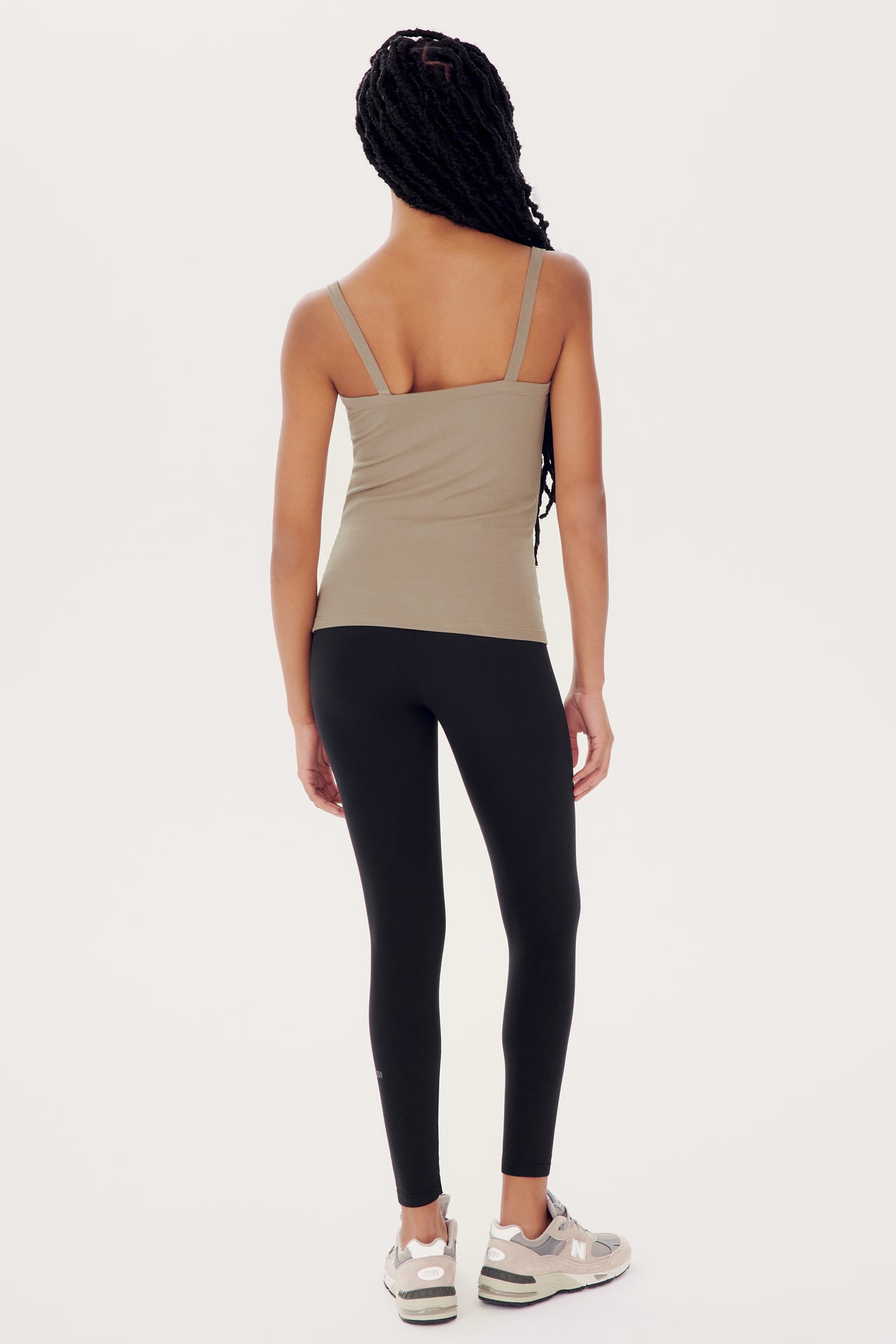 A woman standing with her back to the camera, wearing black leggings, a Romy Rib Tank in Latte by SPLITS59 with a square neckline, and white sneakers.