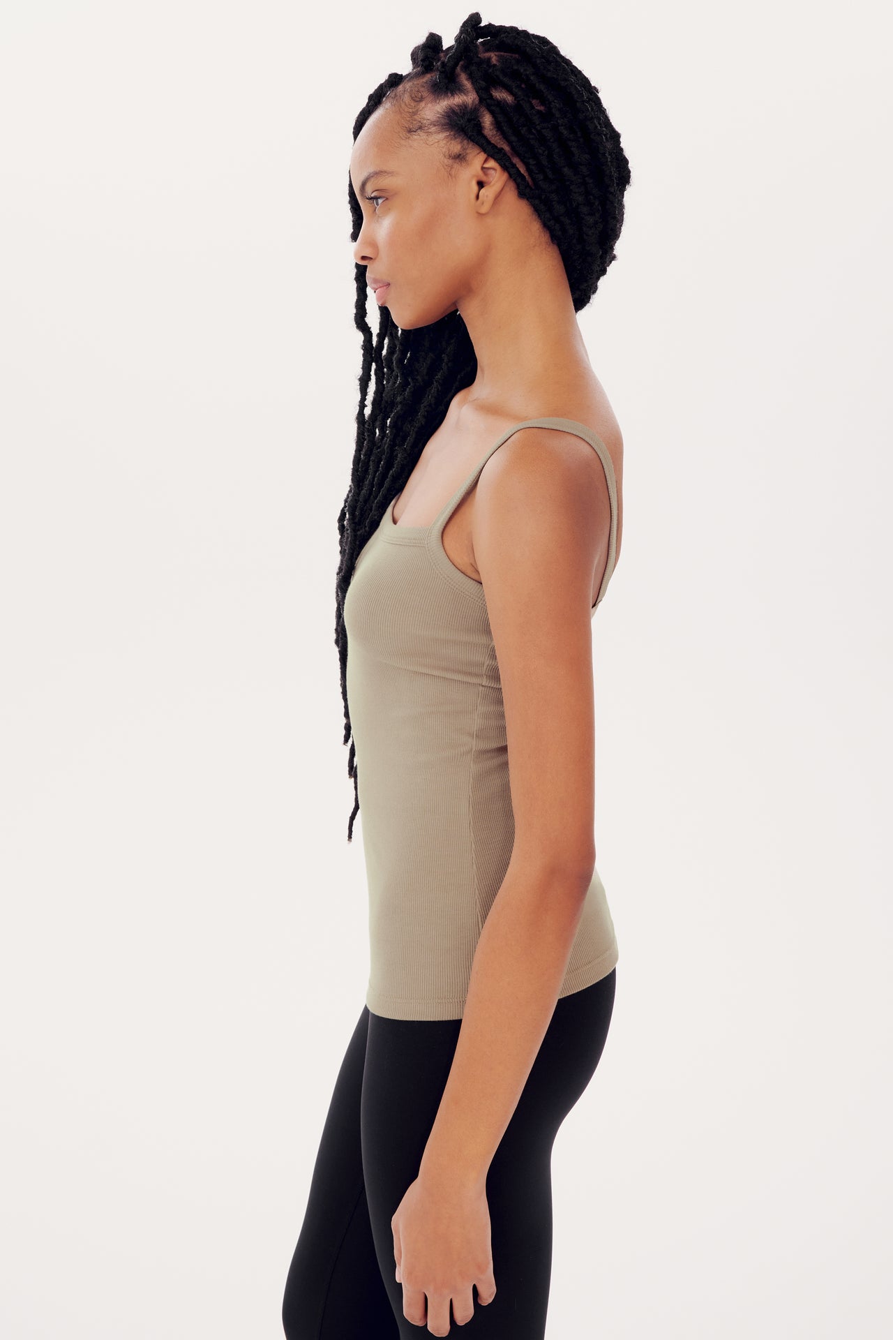 Side profile of a young woman with long braids wearing a Romy Rib Tank in Latte by SPLITS59 with a square neckline and leggings, standing against a white background.