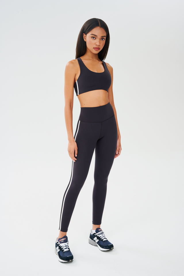 Full side view of girl wearing dark grey sports bra with a thin white and black stripes down the side and dark grey leggings with dark blue shoes