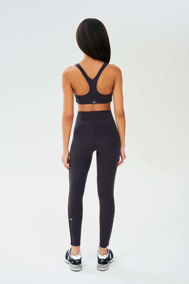 Full back view of girl wearing dark grey sports bra with a thin white and black stripes down the side and dark grey leggings with dark blue shoes