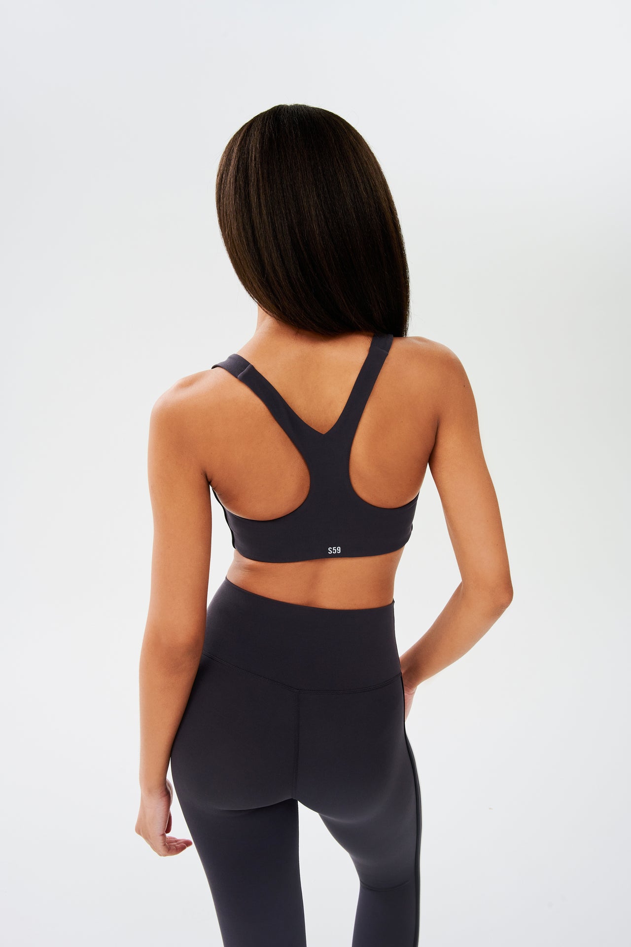Back view of girl wearing dark grey sports bra with a thin white and black stripes down the side and dark grey leggings 