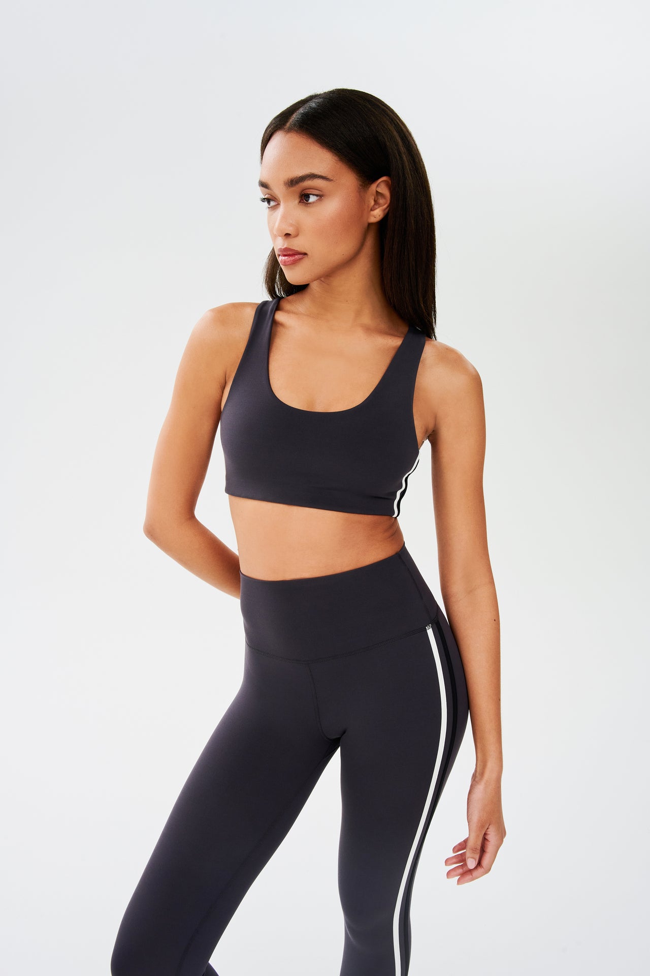 Side view of girl wearing dark grey sports bra with a thin white and black stripes down the side and dark grey leggings 