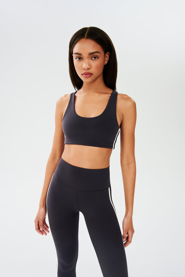 Front view of girl wearing dark grey sports bra with a thin white and black stripes down the side and dark grey leggings 