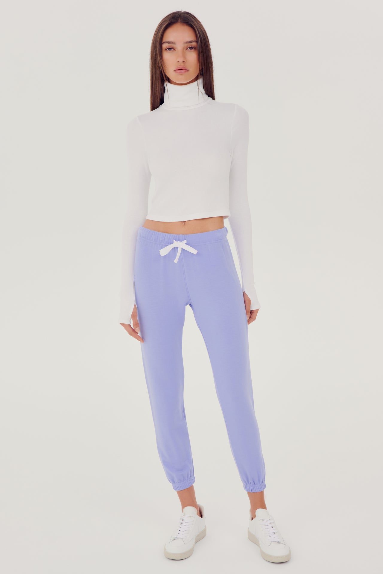 Full front side view of woman with straight dark brown hair wearing light purple sweatpant jogger with white drawstring and white cropped turtleneck long sleeve paired with white shoes 