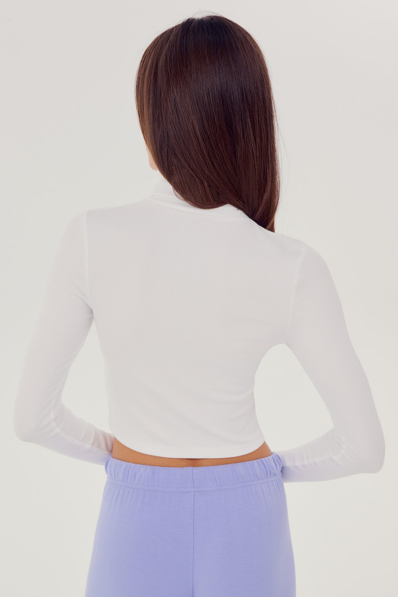 The back view of a woman wearing a SPLITS59 Jackson Rib Cropped Turtleneck in White and high waist purple leggings.