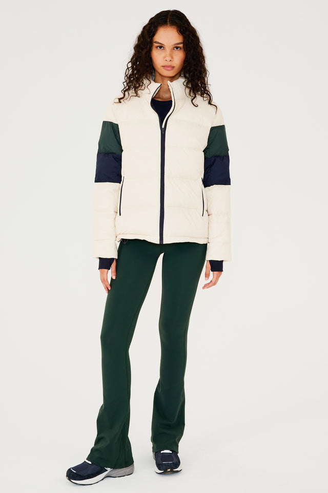 Full front view of girl wearing white puffer jacket with blue and green stripes on arms and sleeve cuffs with thumb holes and dark green flared leggings with dark blue shoes