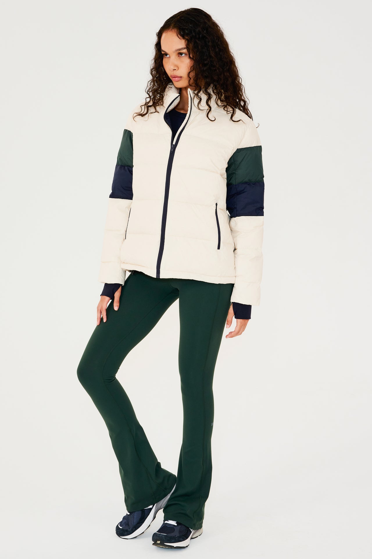 Full side view of girl wearing white puffer jacket with blue and green stripes on arms and sleeve cuffs with thumb holes and dark green flared leggings with dark blue shoes