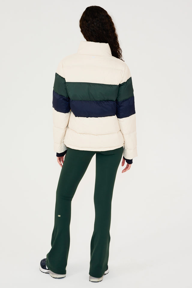 Full back view of girl wearing white puffer jacket with blue and green stripes on arms and sleeve cuffs with thumb holes and dark green flared leggings with dark blue shoes