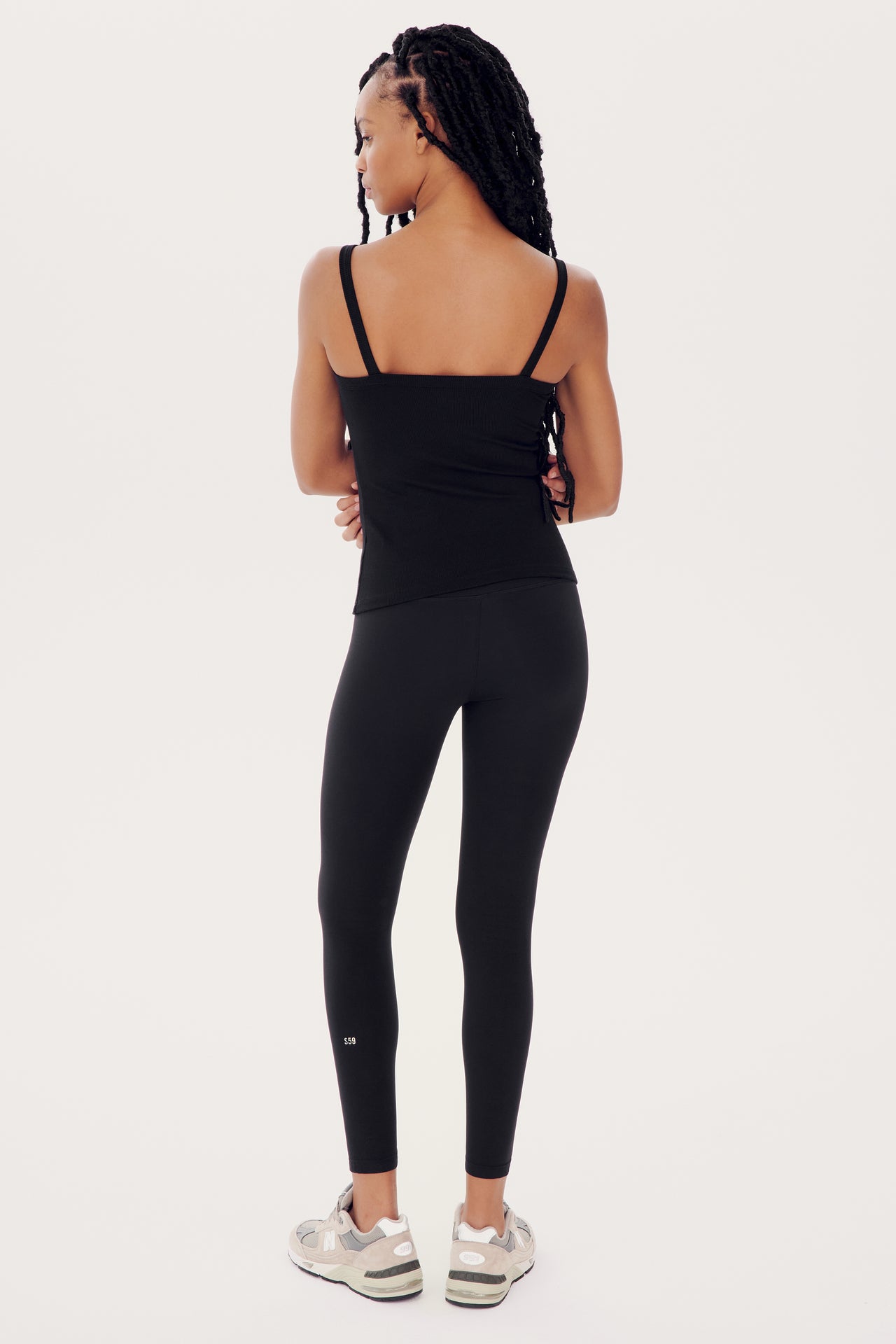 A woman standing with her back to the camera, wearing black leggings and a SPLITS59 Romy Rib Tank - Black, looking to the left, against a white background.